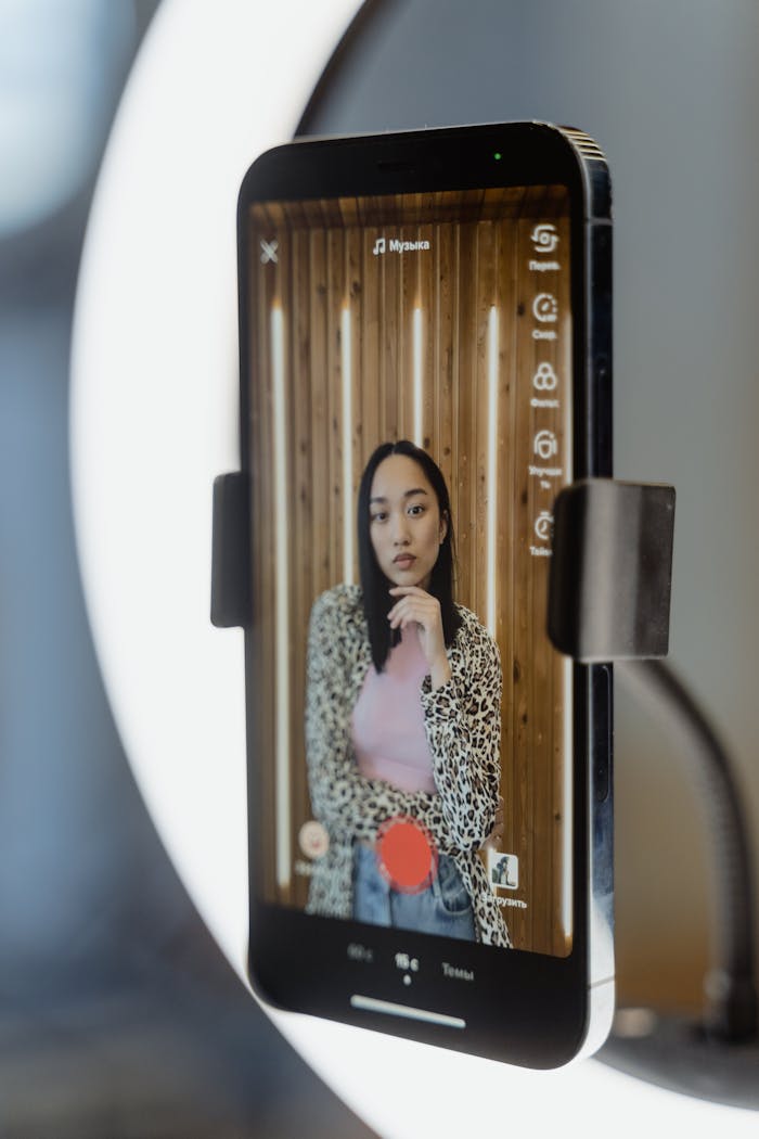 Asian woman recording a TikTok video using a smartphone and ring light indoors.